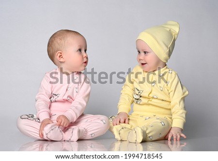 Two baby toddler babies are sitting next to each other on a light studio background. One toddler is dressed in a yellow cotton jumpsuit with a cute hat, the other in a pink sandbag. Children's clothes