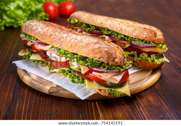 Two baguette sandwiches with salami,\
turkey breast, cheese, lettuce, tomatoes and onion on a cutting\
board. Long subway sandwiches on a dark\
background.