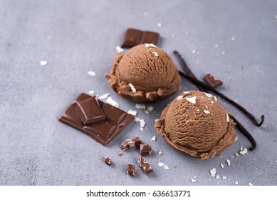 Two balls of chocolate ice cream with a vanilla stick and chunks of chocolate on a dark background
