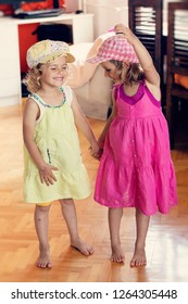 Two Barefoot Cute Twin Sister Girls In Pink And Yellow Dresses And Hats Holding Hands, Standing On Parquet Floor At Home - Shutterstock ID 1264305448