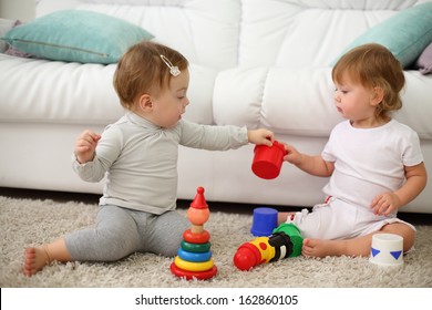 Two barefoot kids sit on carpet and play with colored toys near sofa. Focus on left kid. Shallow depth of field. 