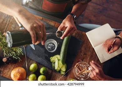 Two Bartenders Experimenting With Creating Cocktail Drinks. Top View Of  Man Pouring Mixture Into A Jigger To Prepare A Cocktail And Another Taking Down Notes.