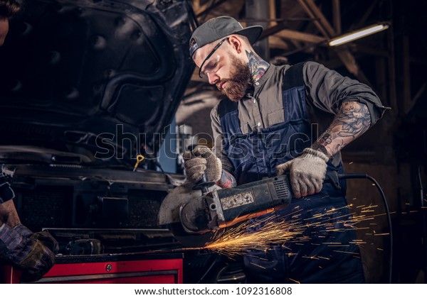 Two bearded auto mechanic in a uniform and safety\
glasses working with an angle grinder while standing against a\
broken car in repair\
garage.