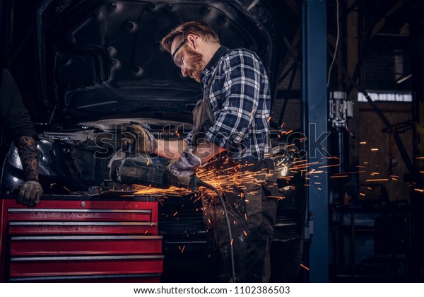 Two bearded auto mechanic in a uniform and\
safety glasses working with an angle grinder while standing against\
a broken car in repair garage.\
