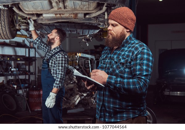 Two bearded brutal mechanics repair a car on a lift in\
the garage. 