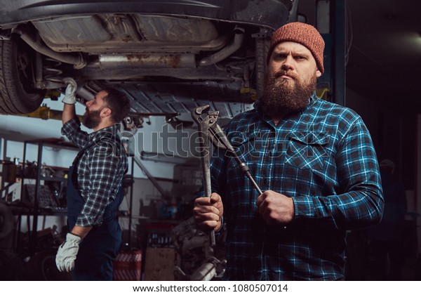 Two bearded brutal mechanics repair a car on a\
lift in a garage.