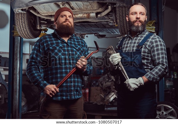 Two bearded brutal mechanics repair a car on a\
lift in a garage.