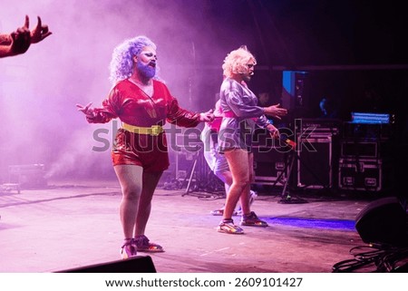 Two bearded drag queens perform on stage, captivating the audience with their bold costumes and energetic movements. The atmosphere is filled with lively music and a sense of celebration.