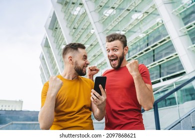 Two Bearded Guys Showing Celebrating Victory In Online Lottery. Friends Being Happy Winning A Bet In Online Sport Gambling Application With Football Stadium On The Background.