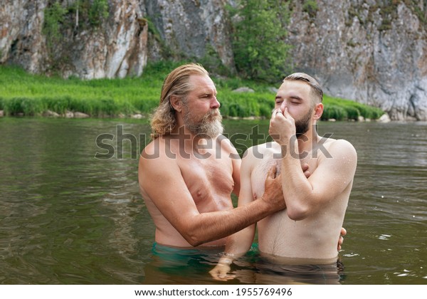 Two bearded men stand in waist-deep water young\
man with eyes closed and nose pinched by hand listens of older man\
who holds hands on his chest and back. learn to dive. Ceremony of\
Christening outdoor