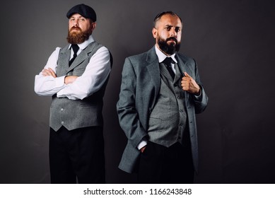 Two Bearded Men In Strict Suits Standing One Near One And Looking On Camera On Black