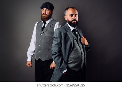 Two Bearded Men In Strict Suits Standing One Near One And Looking On Camera On Black