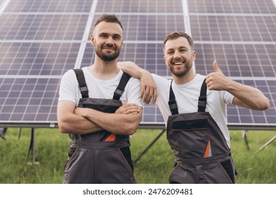 Two bearded men technicians professionals servicing solar farm standing with crossed arms and showing thumbs up near solar panels
