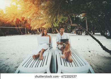 Two beautiful adult people on resort beach chairs: smiling handsome man watching movie via digital tablet and charming woman working on her modern laptop during their vocation on warm sunny evening