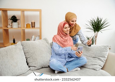 Two beautiful Arab women look at their smartphones and share news. Woman found shopping site. Online shopping concept at home. Girls in hijabs have a pleasant homely atmosphere