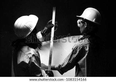 Two Beautiful Asian Architect Engineer women in hard hat, protector glasses goggle mask ear muff, ruler chart blueprint, studio lighting dark smoke background monotone copy space, concept women can do