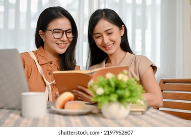 Two Beautiful Asian Female College Students Reading A Book Together, Working On Their Project Together In The Coffee Shop.