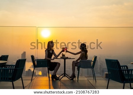 Two beautiful Asian woman friends meeting and dinner party together at skyscraper rooftop restaurant in metropolis at summer sunset. Female friend enjoy outdoor lifestyle activity in the city at night