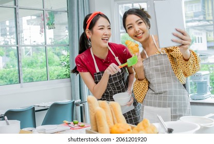 Two Beautiful Asian Women Cooking Food, Making Bread For Breakfast And Bakery While Using Tablet, Streaming Live Video Clip Or Taking Selfie Photo Together With Happiness And Fun In Kitchen At Home