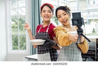 Two Beautiful Asian Women Cooking Food, Making Bread For Breakfast And Bakery While Using Tablet, Streaming Live Video Clip Or Taking Selfie Photo Together With Happiness And Fun In Kitchen At Home