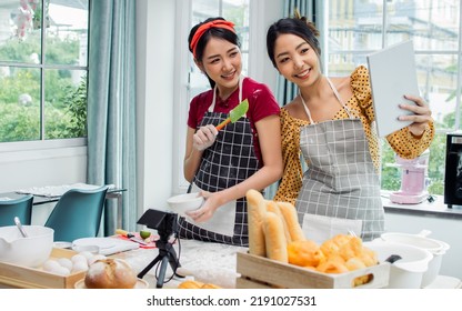 Two Beautiful Asian Women Cooking Food, Making Bread For Breakfast And Bakery While Using Tablet, Streaming Live Video Clip Or Taking Selfie Photo Together With Happiness And Fun In Kitchen At Home