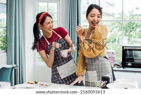 Two beautiful Asian women cooking food, making bread for breakfast and bakery, teasing, playing together with happiness, fun while standing in kitchen at home on weekend. Hobby and Lifestyle Concept