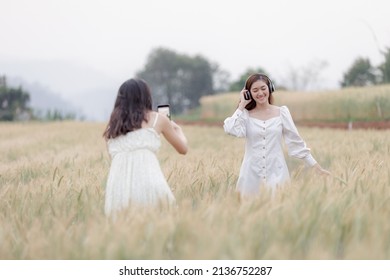 two beautiful asian women girl in while dress taking photo with smartphone and listen to music wireless headphone in agriculture demonstration plots, barley rice fields at chiang mai Thailand,

