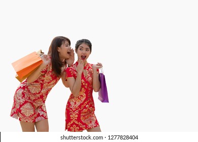 Two Beautiful Asian Women, Hold Shopping Bags, One Whisper In Friend's Ear Who Has Excited Face, Both Are Enjoy Celebrating Chinese New Year, Dress In Red Qipao/cheongsam.Isolated On White Background.