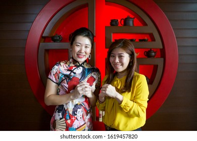 Two beautiful Asian young female greet with both hands to celebrate Chinese New Year. Asia culture .Ladies with qipao and yellow top.Teapots and cups on background shelves.Brown wooden wall.