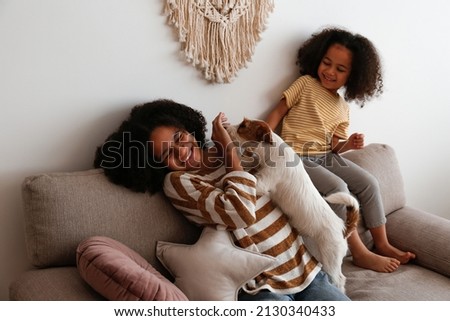 Two beautiful black girls of different age playing with their adorable wire haired Jack Russel terrier puppy at home. Loving sisters with rough coated pup having fun. Background, close up, copy space.