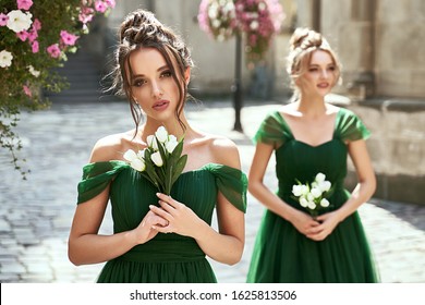 Two beautiful bridesmaids girls blonde and brunette ladies wearing elegant full length off-the-shoulder green chiffon bridesmaid dress and holding flower bouquets. European old town location for