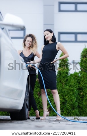 Two beautiful businesswomen in black outfut posing next to an electric car. Girls plugging blue cable in power charging socket.
