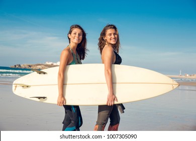 Two Beautiful Female Friends At The Beach Holding A Surfboard And Smiling