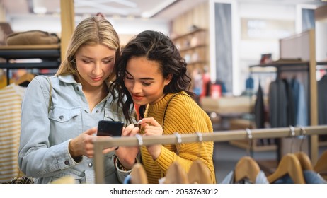 Two Beautiful Female Friends Shopping In Clothing Store, Using Smartphone, Browsing Online, Comparing On Internet. Customers Choosing Clothes. Fashionable Shop, Colorful Brands, Sustainable Designs.