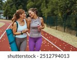 Two beautiful fitness female friends in sportswear holding gym bag, yoga mat and water bottle talking in the park. Sporty girl ready for training. Two female friends spending time together