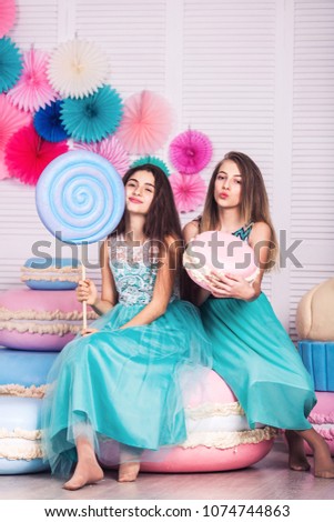 Two beautiful girls in blue dresses hold in their hands huge candy and macaroon in studio with decor of macaroons.