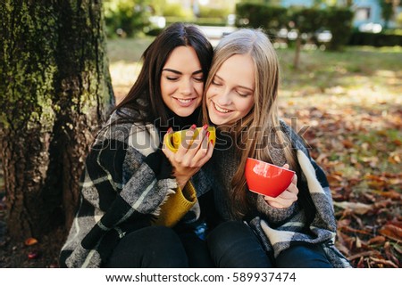 two beautiful girls bundled up plaid and holds cupsin hands in the park, posing for the camera