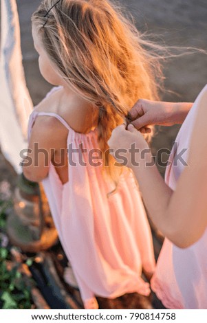 Two beautiful girls in light dresses float on a lake on a raft with a sail and braid each other pigtails at sunset