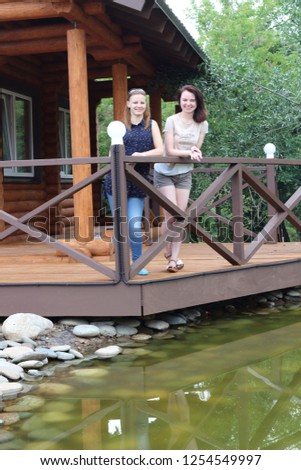 Two beautiful girls in on the background of a wooden cottage and an artificial pond in the park. Nice girl in the park in the countryside.