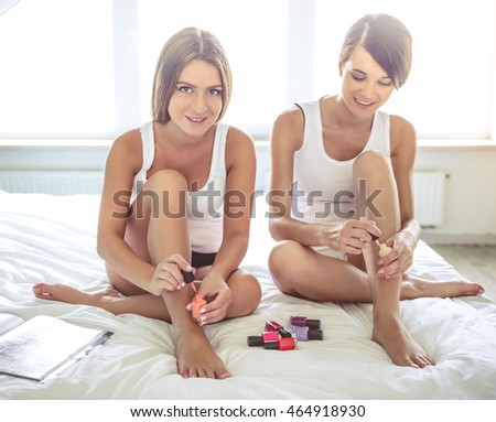 Two beautiful girls are using nail polish, talking and smiling while doing pedicure on bed at home. One girl is looking at camera
