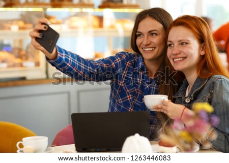 Two beautiful girls wearing in casual clothing, taking self portrait at smart phone. Friends smiling and posing, sitting at bakery shop. Redhead girl holding cup of coffee. Concept of friendship.