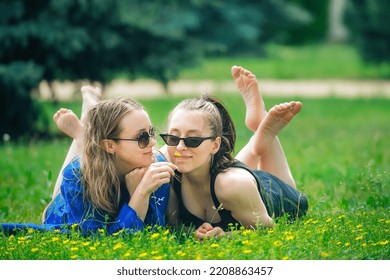 Two Beautiful Happy Girls Girlfriends Lie On Bright Green Grass On The Lawn, Chat Merrily And Sniff Yellow Flowers