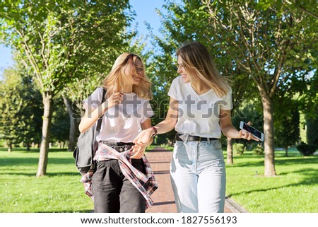 Two beautiful happy girls teenagers 17, 18 years old walking together in park, girls laughing talking having fun on sunny summer day