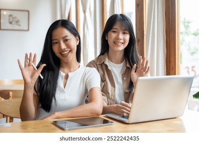 Two beautiful, happy young Asian female friends are sitting at a table in a minimalist coffee shop, smiling and waving at the camera, with a digital tablet and a laptop placed on the table. - Powered by Shutterstock