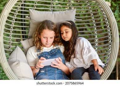 Two Beautiful Little Girl Using A Smart-phone, Sitting Together In Comfy Hanging Egg Chair In A Garden.