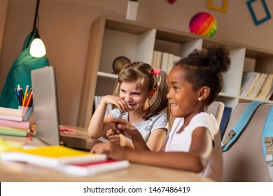 Two beautiful little girls sitting at a desk, watching cartoons on a laptop computer and having fun