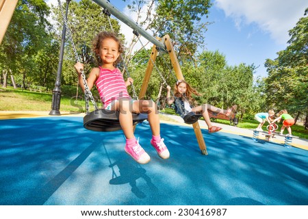 Two beautiful little girls swinging on swings on playground with smile on sunny summer day