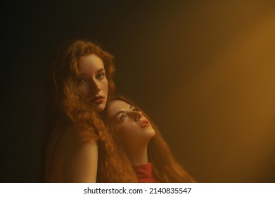 Two beautiful redhead freckled women with long natural curly hair, red lips makeup posing in darkness and warm light. Studio art portrait. Copy, empty space for text