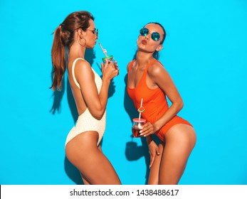 Two beautiful sexy smiling hipster women in summer colorful swimwear bathing suits.Trendy girls in sunglasses.Going crazy.Funny models isolated on blue.Drinking fresh cocktail smoozy drink
