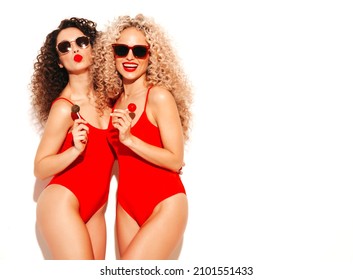 Two beautiful smiling hipster women in red swimwear bathing suits.Trendy models with curls hairstyle in studio. Hot female posing near white wall in sunglasses.Holding lollipop. Isolated on white